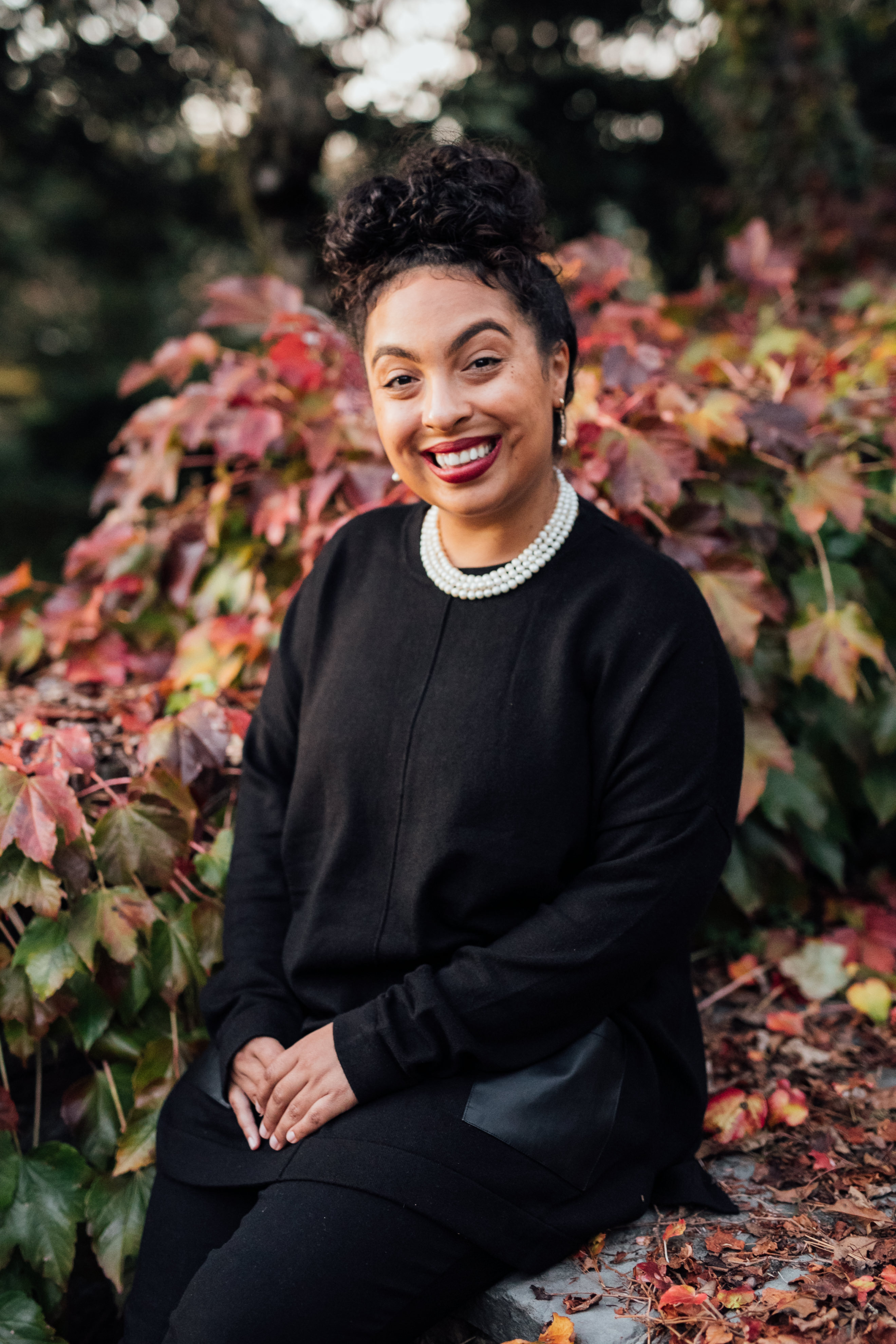 Photo of Julia, a Black woman with curly brown hair that is gathered in a bun on top of her head. She is smiling at the camera and wearing a black shirt.